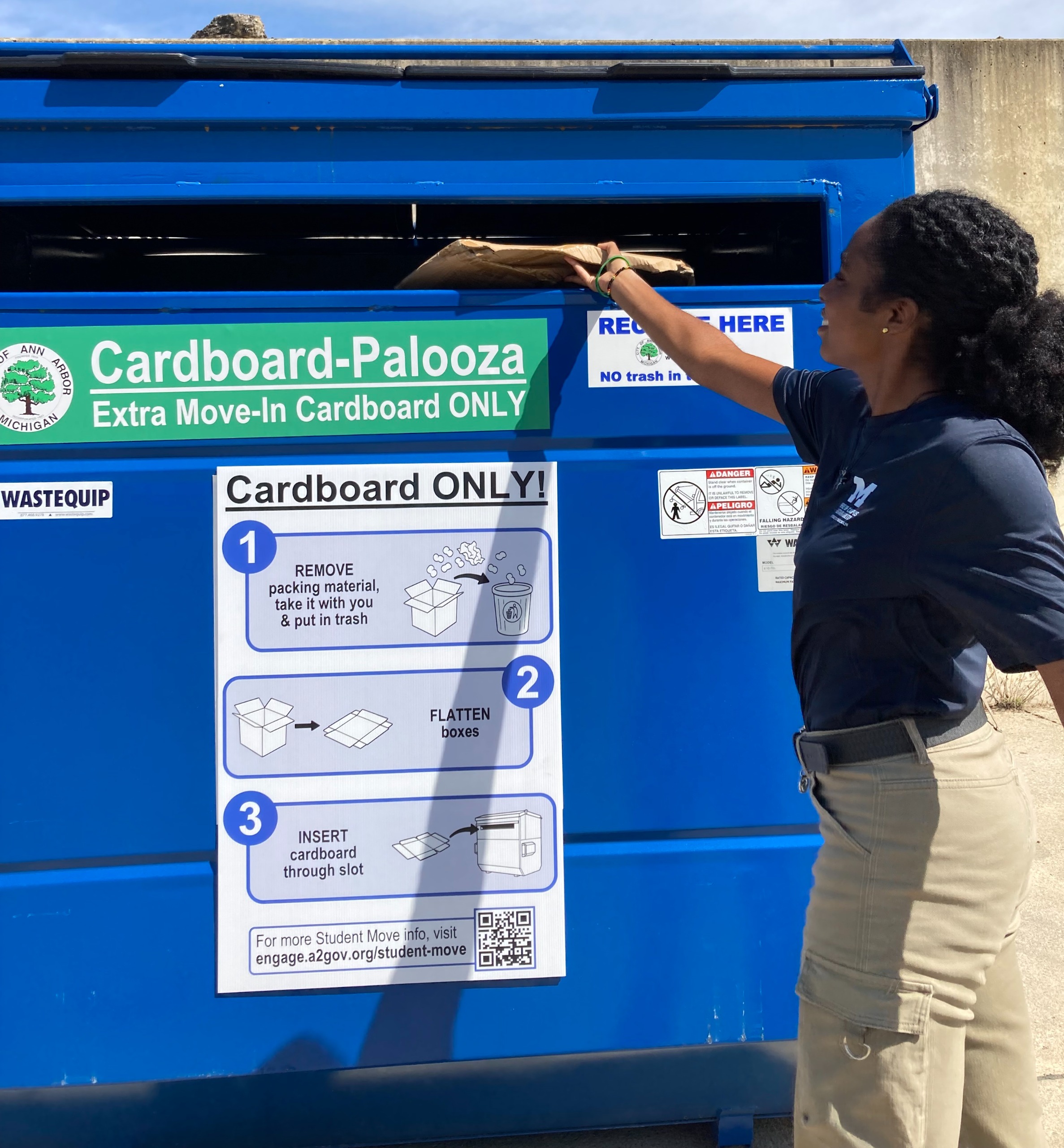 Woman putting cardboard box through slot of cardboard dumpster. 